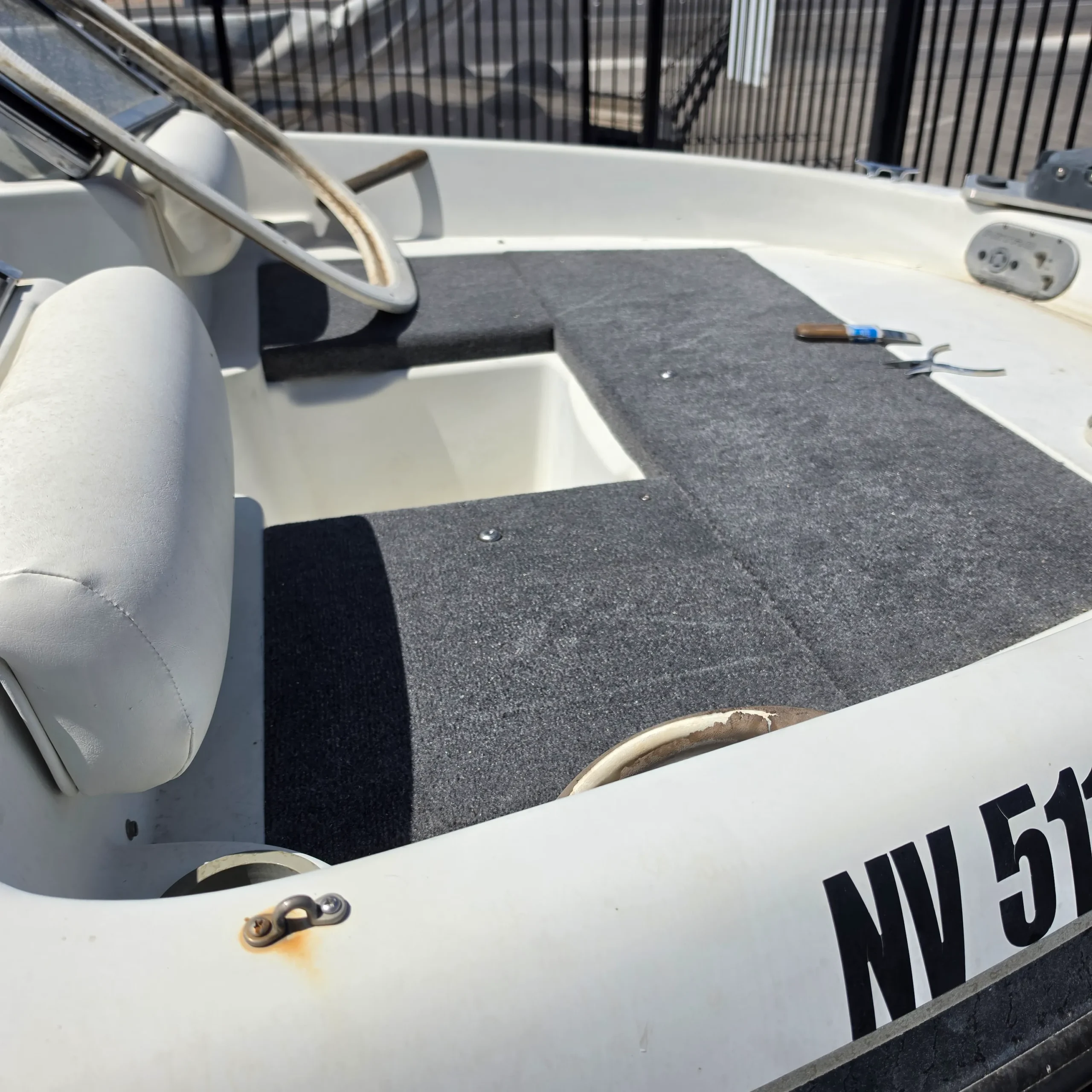 Boat with a steering wheel and a seat in it, surrounded by a railing and a road in the background