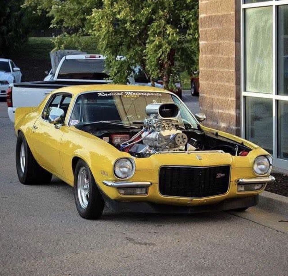 Yellow 1970 RS/SS parked in front of a building with glass windows, surrounded by trees and grass